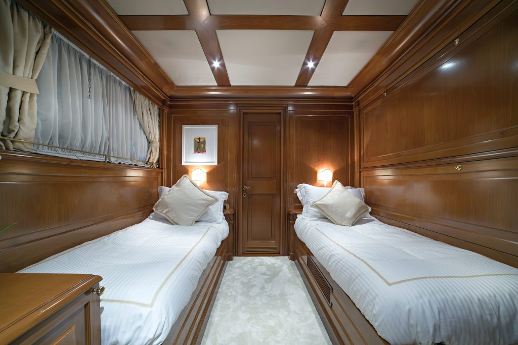 Luxurious cabin room with polished wooden walls and ceiling, two single beds with white bedding, a closed door at the center, wall lamps above each bed, and a framed picture—perfect for your next yacht charter.