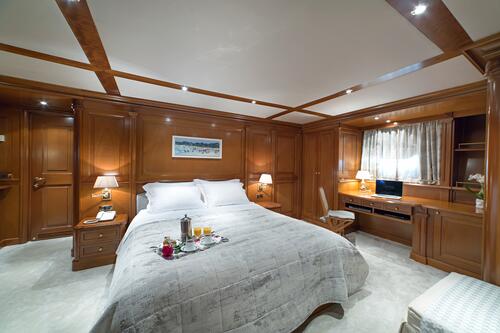 A luxurious bedroom with wood-paneled walls and a large bed with white bedding, perfect for relaxing during your yacht charter. Bedside tables with lamps, a desk with a computer, and a breakfast tray create an inviting space near the window curtains.