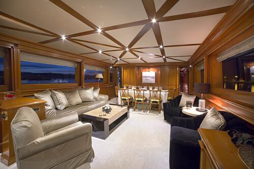 Luxurious yacht interior featuring plush beige sofas, black armchairs, a modern coffee table, wooden paneling, large windows, and a dining area set for dinner—perfect for those seeking premium yachts for charter—all warmly lit with ceiling spotlights.