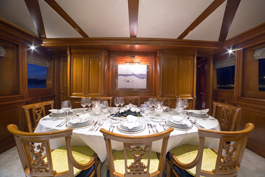 A luxurious dining room with polished wooden walls and ceiling beams, typical of yachts for charter, features a round table elegantly set with plates, glassware, and silverware. Ornate wooden chairs surround the table, and a framed photo decorates the wall.