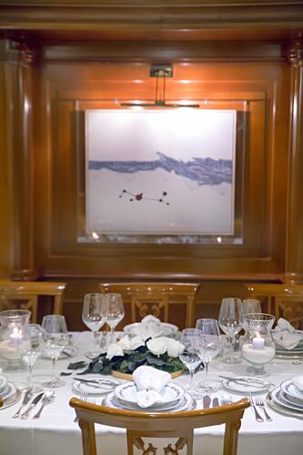 A formal dining table set with white plates, wine glasses, candles, and a floral centerpiece in an elegant wood-paneled room—perfect for a refined yacht charter experience, complete with a framed abstract painting on the wall.