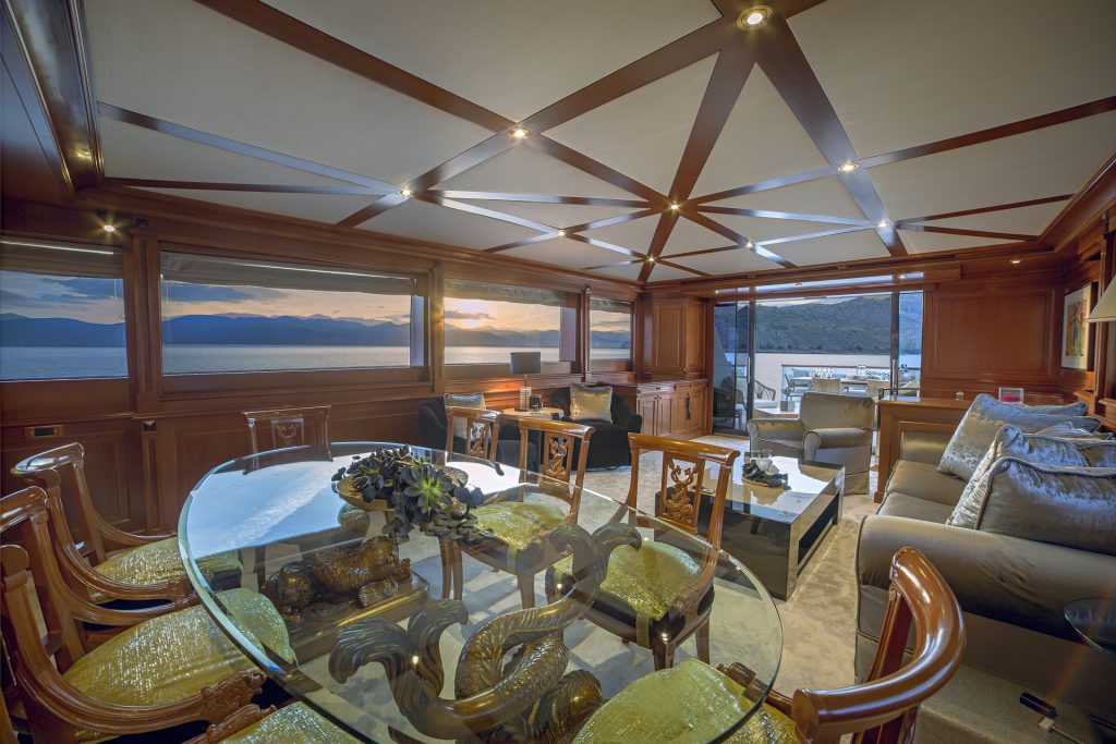 Spacious yacht interior with wood paneling, a glass dining table, cushioned chairs, and sofas. Perfect for a memorable yacht charter, large windows reveal a scenic ocean sunset while the deck with outdoor seating awaits in the background.