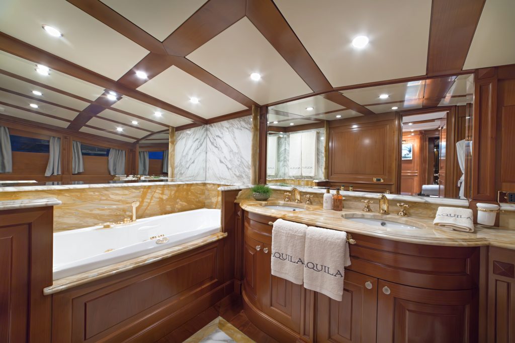Luxurious bathroom with rich wood paneling, marble surfaces, a large bathtub, double sinks, and a towel labeled QUILA QUILA—exquisite details you’d find on exclusive yachts for charter.