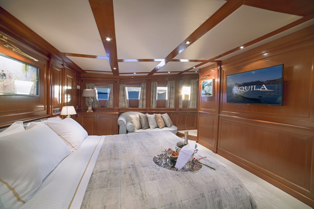 Luxurious yacht bedroom with wood-paneled walls, a large bed with white linens, cozy sofa, and wall-mounted TV displaying AQUILA. Perfect for a relaxing yachts for charter experience, with sea views and a tray of wine glasses on the bed.