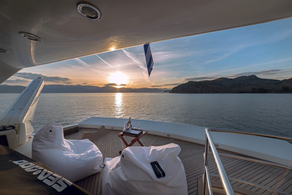 Luxury yacht charter deck at sunset with two white bean bag chairs, a small table holding wine and glasses, calm sea, mountains, and a colorful sky in the background. Peaceful and relaxing atmosphere awaits your next yacht charter escape.