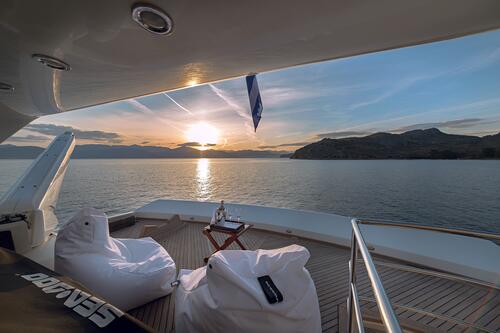Luxury yacht charter deck at sunset with two white bean bag chairs, a small table holding wine and glasses, calm sea, mountains, and a colorful sky in the background. Peaceful and relaxing atmosphere awaits your next yacht charter escape.