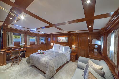 A spacious, luxurious yacht charter bedroom with wood-paneled walls and ceiling, a large bed with gray bedding, a sofa, desk, lamps, and windows allowing ample natural light. A painting hangs above the bed.