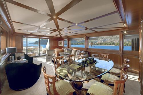 Luxurious yacht interior with wood paneling, a glass dining table, elegant chairs, plush seating, large windows showing a blue sea and rocky coastline—perfect for those seeking an unforgettable yacht charter experience.