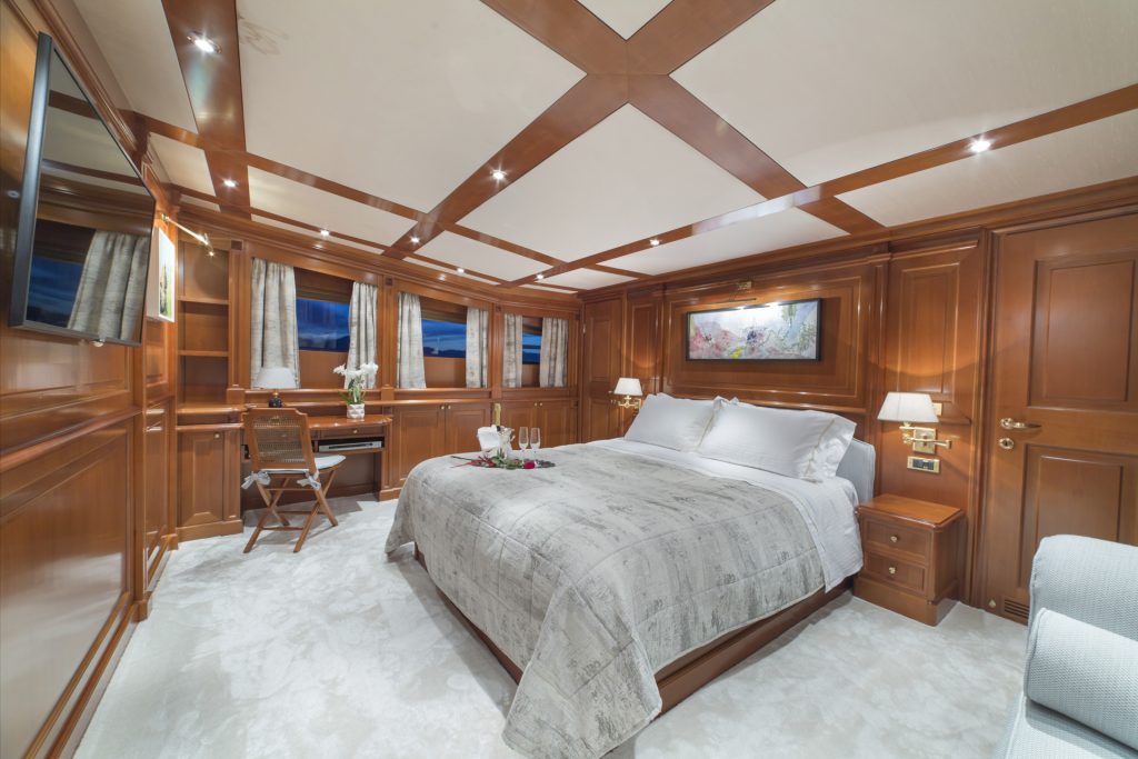 Luxurious yacht bedroom with wood-paneled walls, a large bed with a gray patterned comforter, wall-mounted TV, desk and chair, windows, and soft lighting. Perfect for relaxing on your next yacht rental. A painting hangs above the bed.