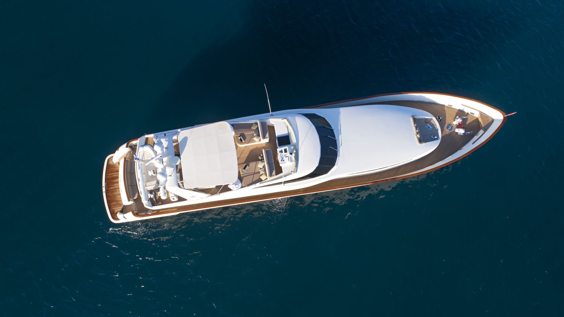 Aerial view of a white luxury yacht with wooden decking floating on calm, deep blue water, showing seating areas and sun loungers—an ideal choice for your next yacht charter.