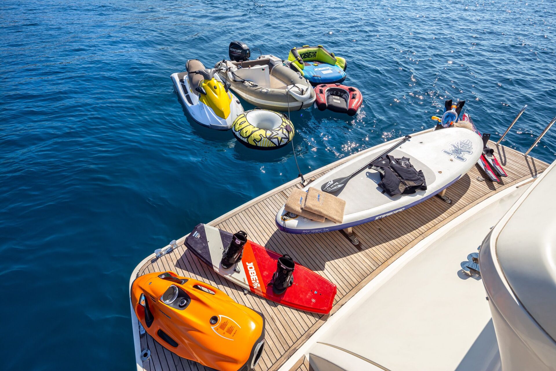 AMORAKI A yacht deck with water sports equipment, including a paddleboard, wakeboard, orange underwater scooter, and fins. Several inflatable boats and floats are anchored nearby in the clear blue sea.