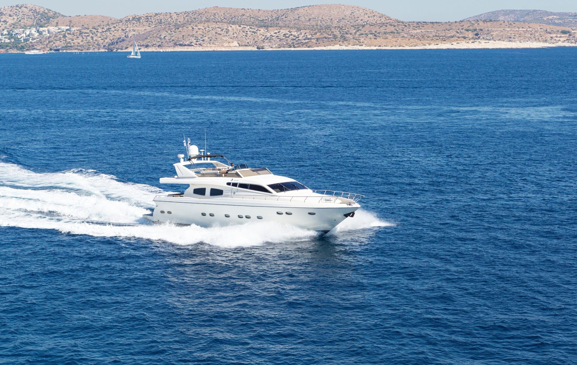 AMORAKI A white luxury yacht speeds across a deep blue sea, leaving a trail of white foam behind. In the background, there are hilly, brown coastlines and a distant sailboat under a clear sky.