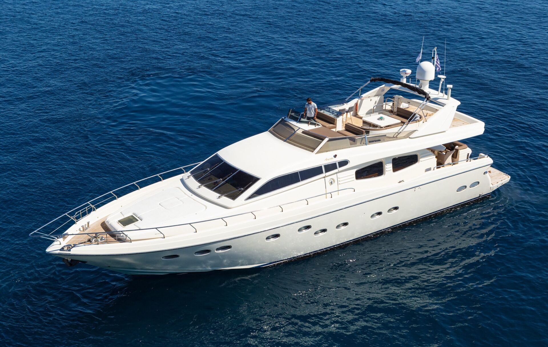 A large white luxury yacht floats on calm blue ocean water, seen from above, with a person standing on the upper deck.