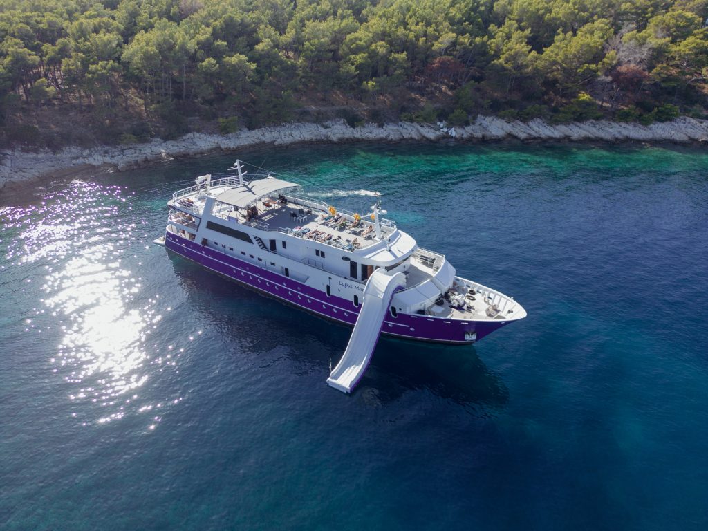 A large yacht with a white and purple exterior is anchored near a forested shoreline in clear blue water. A slide extends from the upper deck into the sea. Sunlight glistens on the water’s surface.