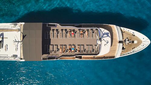 Aerial view of a yacht anchored in bright blue water, with people sunbathing on loungers on the upper deck and a shaded area covering part of the deck.