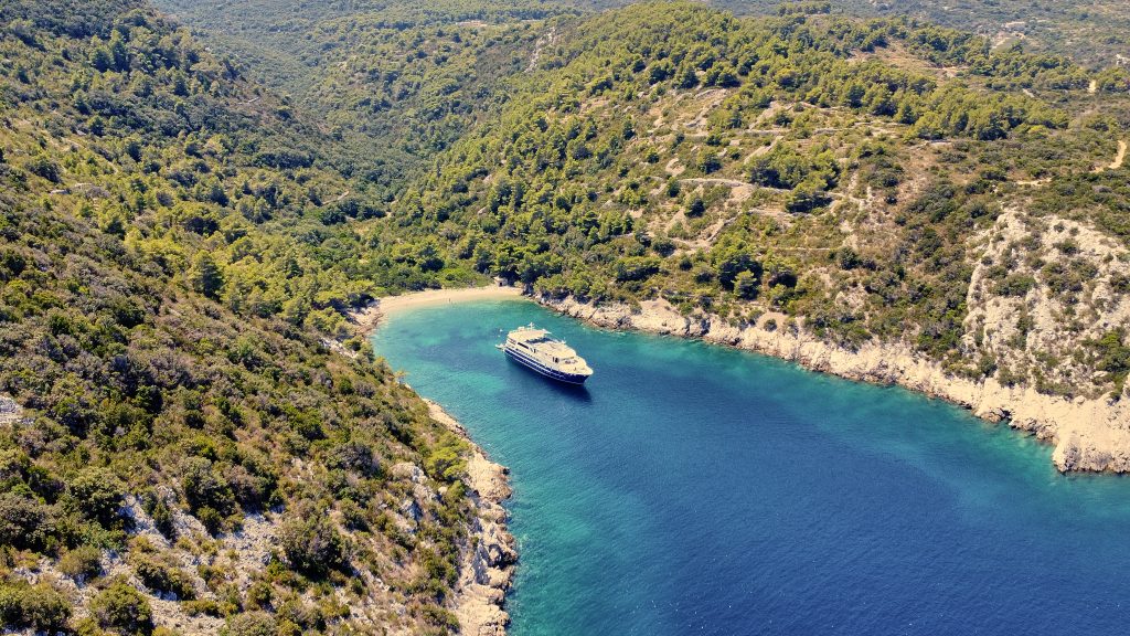 A yacht is anchored in a secluded, turquoise bay surrounded by lush green hills and dense forest under a clear sky.