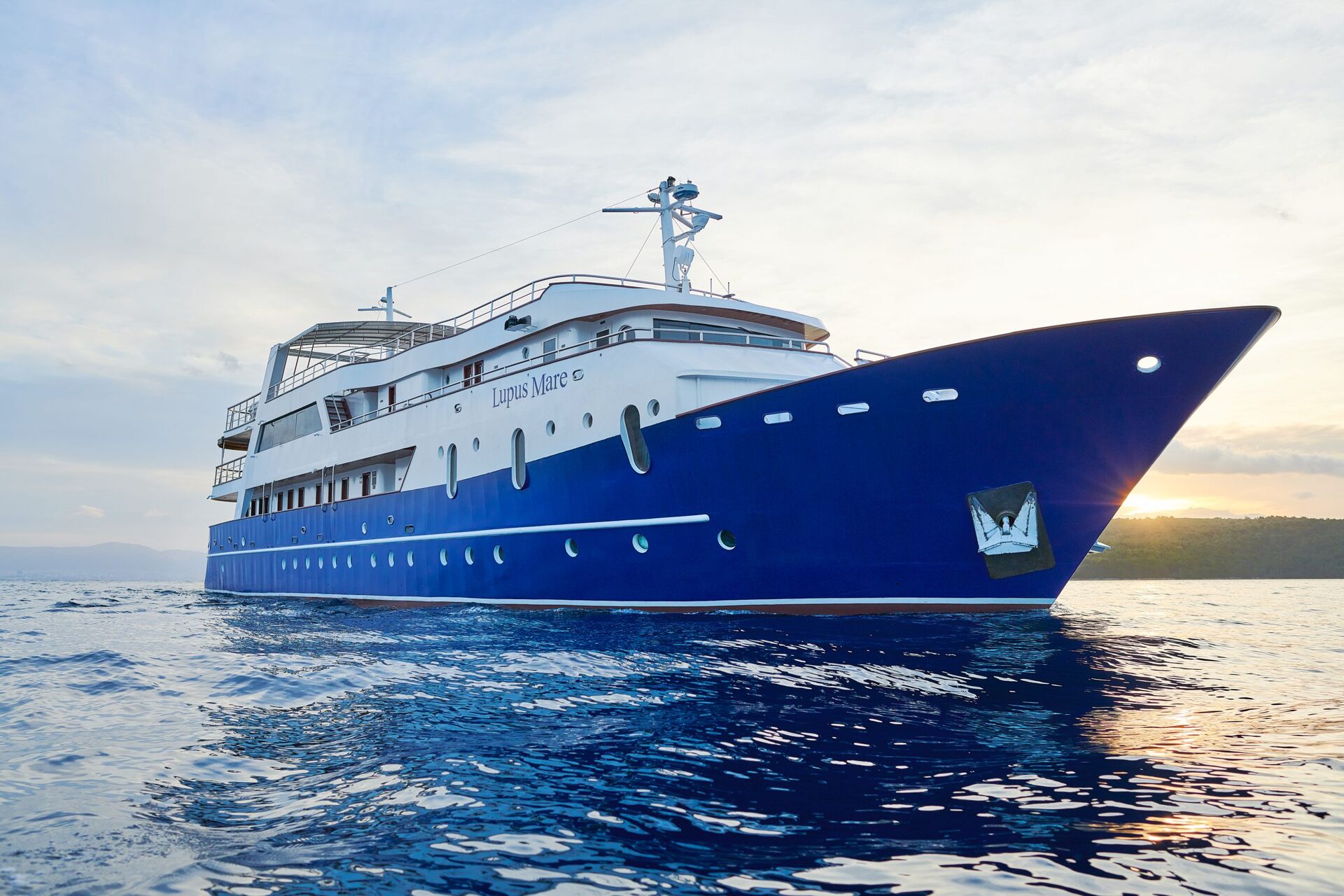 A large, blue and white luxury yacht floats on calm ocean water at sunset, with Lupus Mare written on the side. The sky is partly cloudy, and land is visible in the distance.