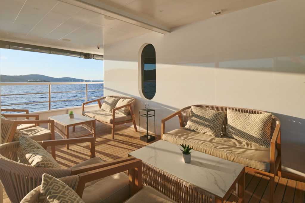 A stylish yacht deck with cushioned wooden chairs and sofas around marble-top tables, overlooking calm blue water and distant hills under soft sunlight.