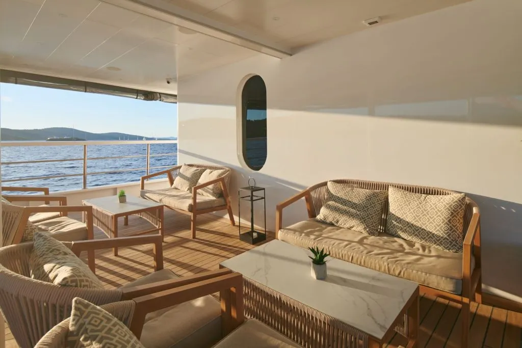 A stylish yacht deck with cushioned wooden chairs and sofas around marble-top tables, overlooking calm blue water and distant hills under soft sunlight.