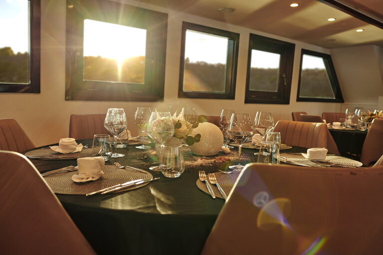 A round dining table set elegantly with glassware, plates, and a white floral centerpiece in a sunlit room with modern brown chairs and several windows letting in sunlight.