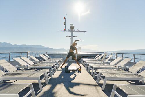 A person exercises with kettlebells on the sun deck of a boat, surrounded by empty lounge chairs, with bright sunlight and mountains in the background.