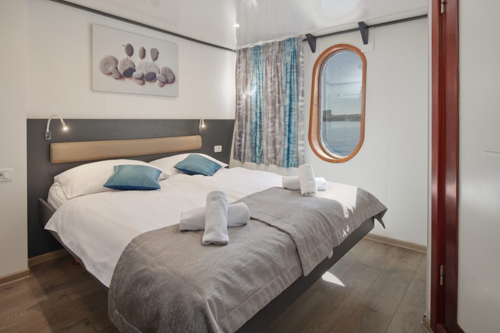 A cozy cabin bedroom with a large bed, blue accent pillows, rolled towels, a round window with a view of water, and modern decor with soft lighting.