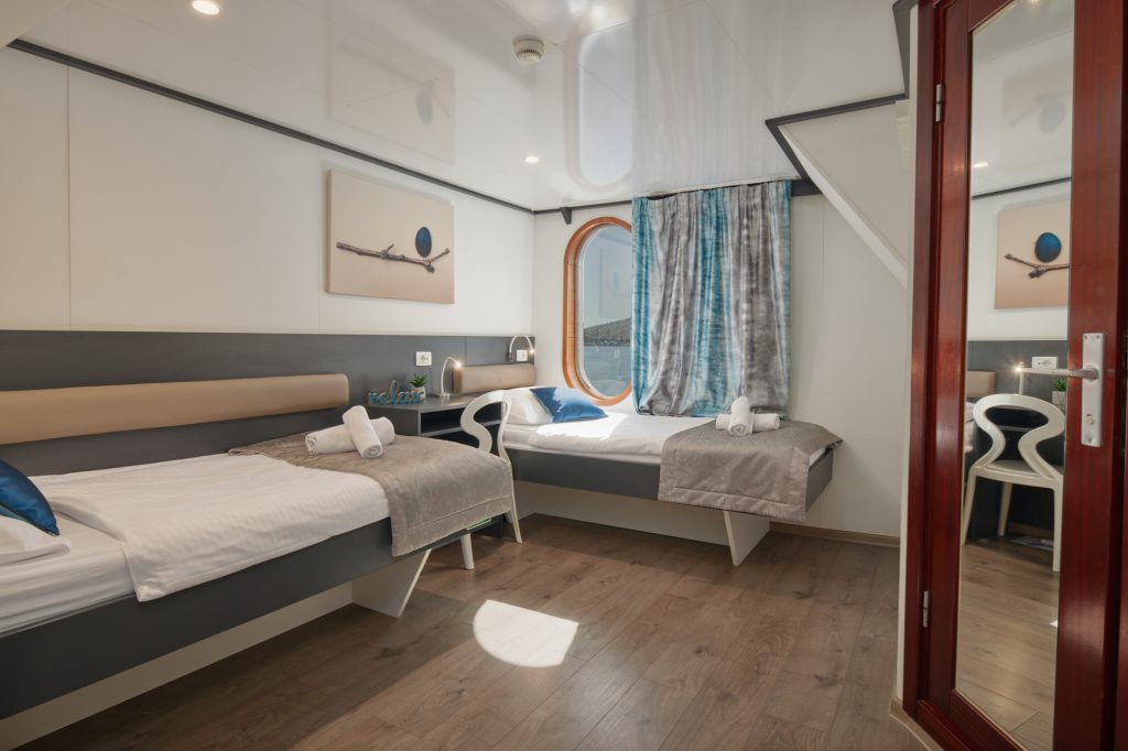 Modern cabin room with two single beds, rolled towels, gray and blue bedding, a round window with curtains, wall art, wood flooring, and a glass door, creating a bright and cozy atmosphere.