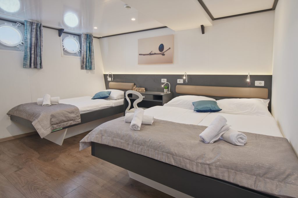A modern cabin room with two double beds, each made with white linens, gray throws, and blue accent pillows. Rolled towels are placed on each bed. The room has wood flooring, porthole windows, and a wall art above the beds.