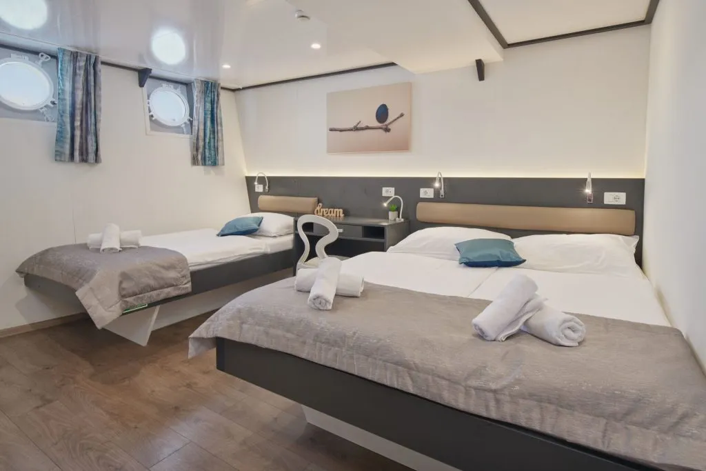 A modern cabin room with two double beds, each made with white linens, gray throws, and blue accent pillows. Rolled towels are placed on each bed. The room has wood flooring, porthole windows, and a wall art above the beds.