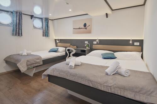 A modern cabin room with two double beds, each made with white linens, gray throws, and blue accent pillows. Rolled towels are placed on each bed. The room has wood flooring, porthole windows, and a wall art above the beds.