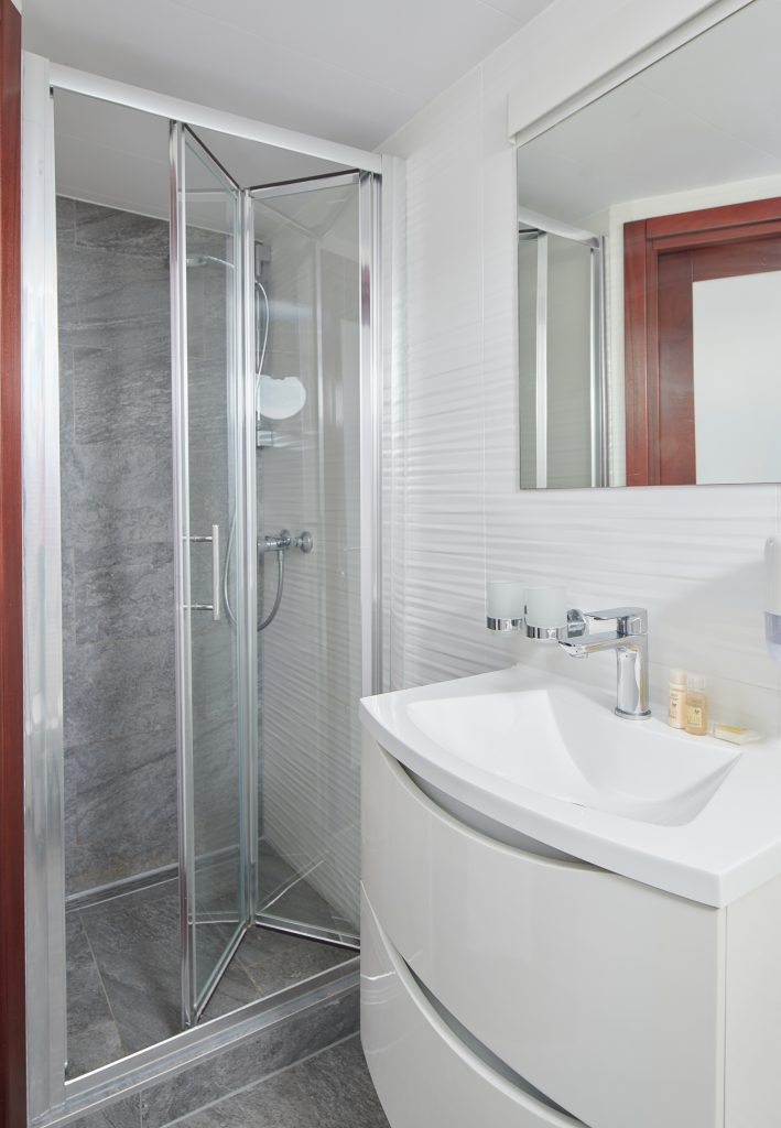 Modern bathroom with a glass shower enclosure, gray tiled walls, a white curved sink vanity with a faucet, and a large mirror above the sink. Toiletries are placed on the sink counter.