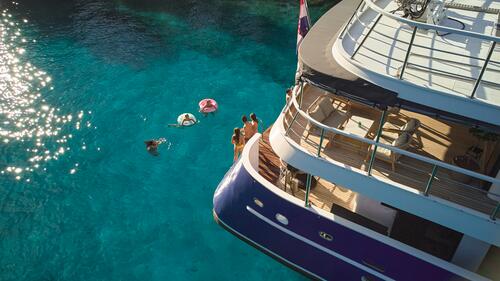Aerial view of people swimming and floating on inflatables in clear blue water next to a large yacht, with sunlight reflecting on the sea.