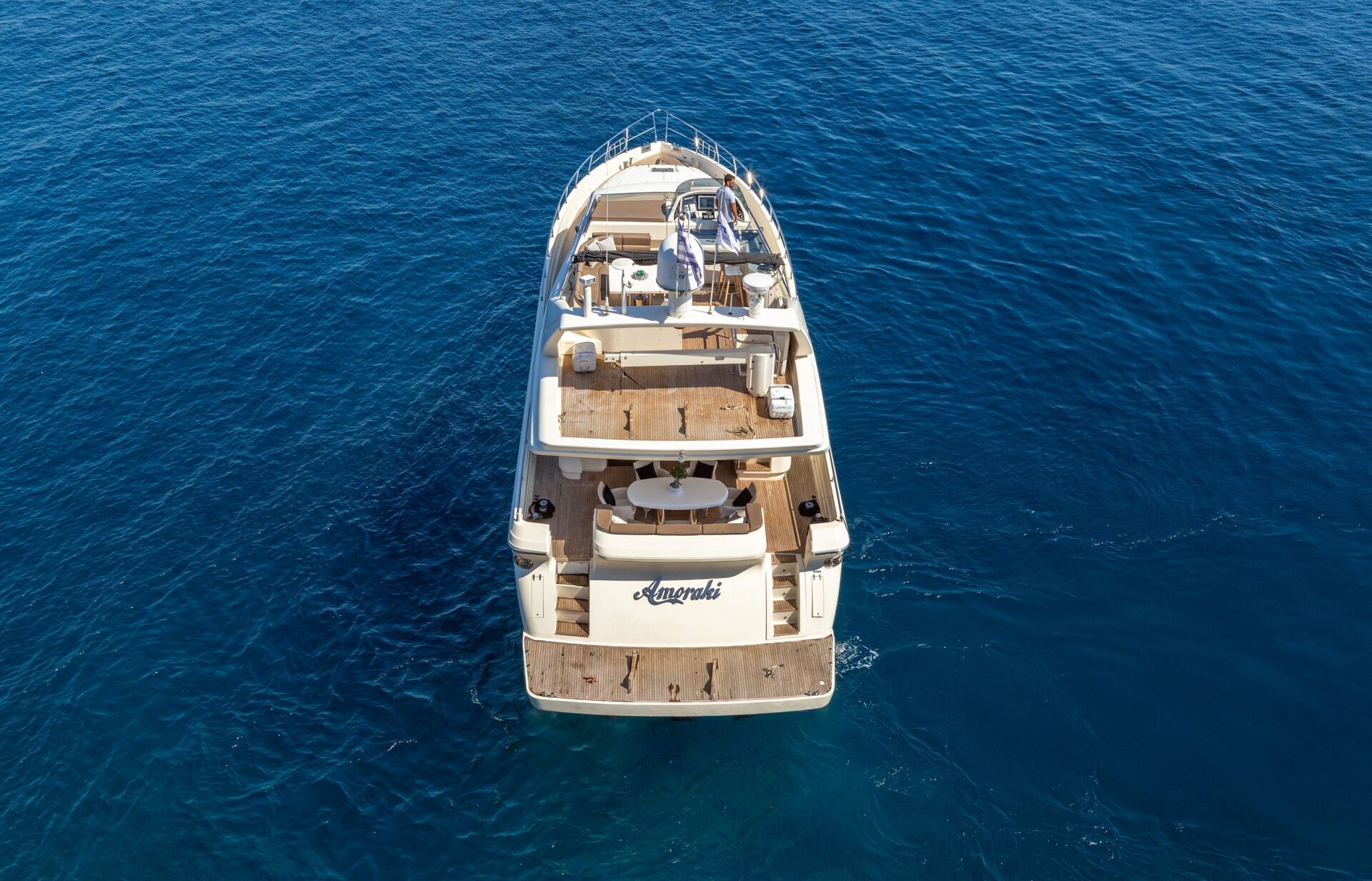 AMORAKI A luxury yacht named Amanda is anchored on calm, deep blue water, viewed from above and behind, showing its decks, seating areas, and sunbathing spaces.