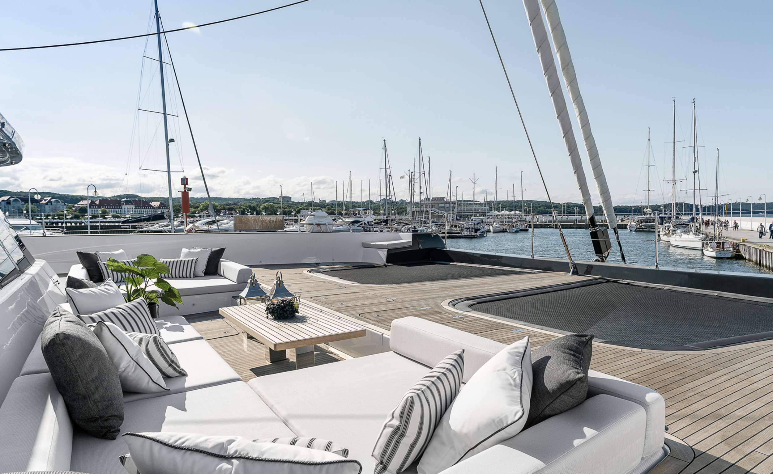 A luxurious yacht charter deck features white cushioned seating with striped pillows, a small table with plants, and views of a marina filled with sailboats under a clear sky.