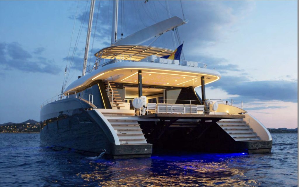 A luxury catamaran yacht for charter is anchored at dusk on calm water, illuminated by blue underwater lights and warm deck lighting, with a scenic coastline and cloudy sky in the background.