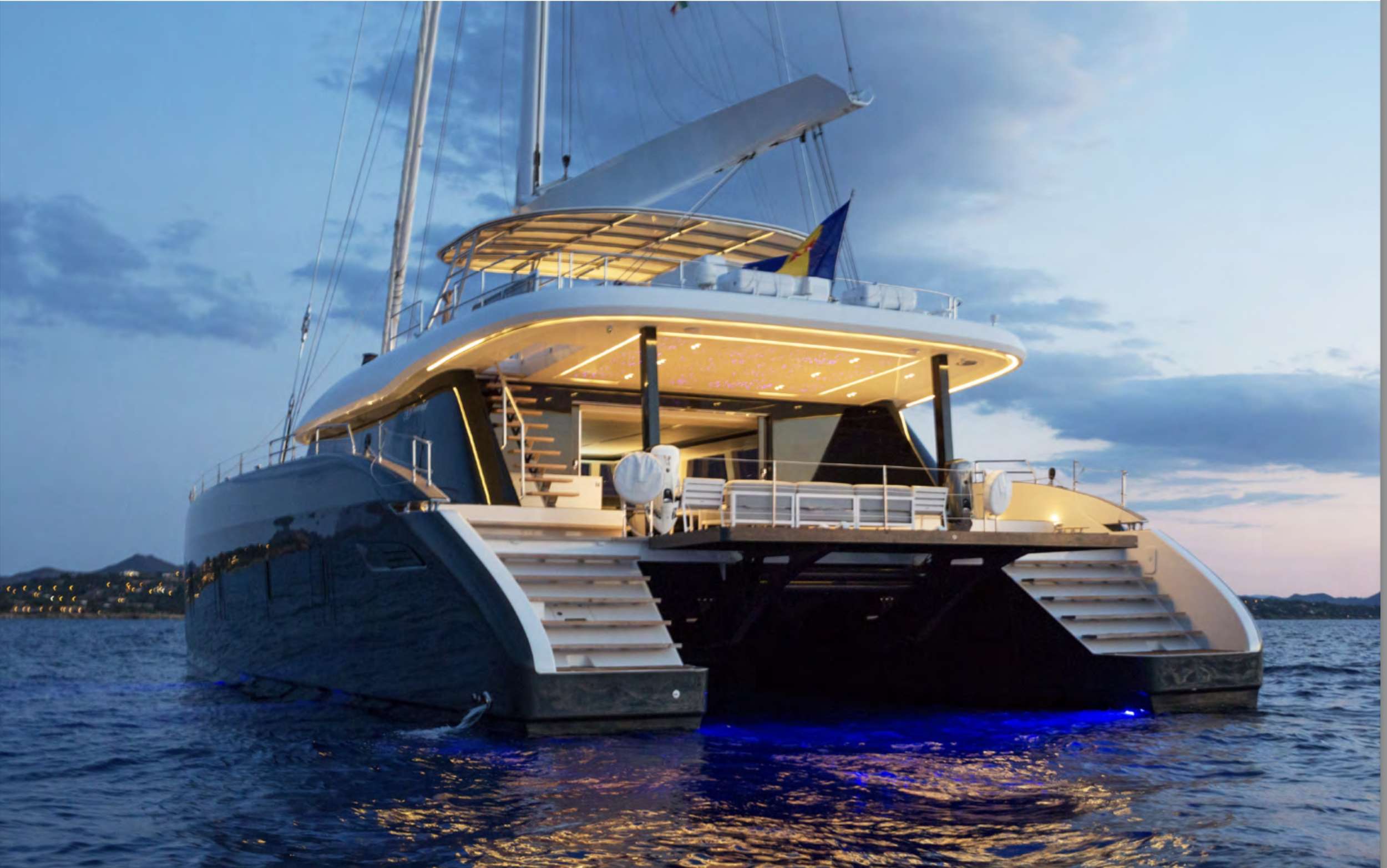 A luxury catamaran yacht for charter is anchored at dusk on calm water, illuminated by blue underwater lights and warm deck lighting, with a scenic coastline and cloudy sky in the background.