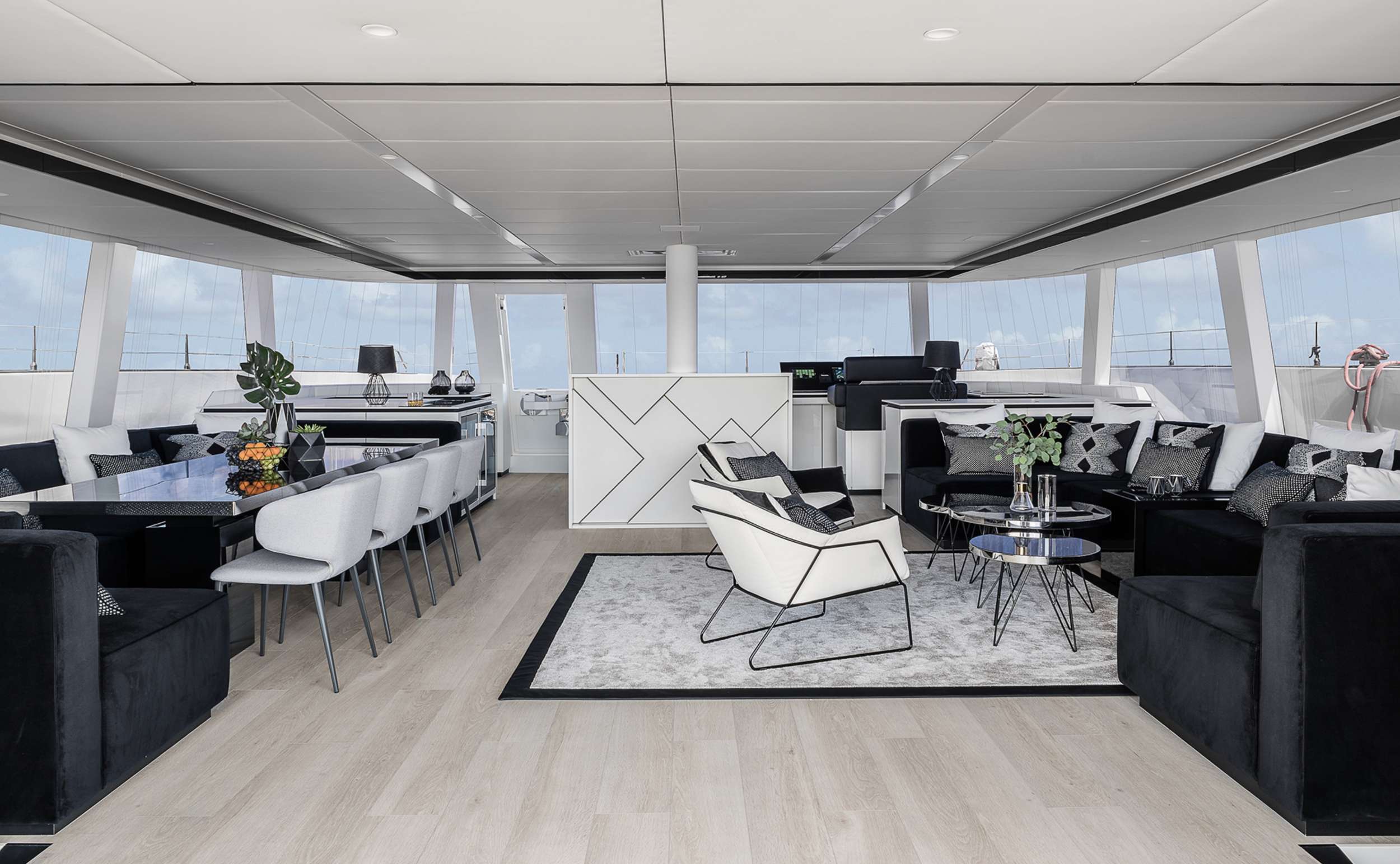 Spacious, modern yacht interior with large windows, black and white furniture—perfect for yacht rental. Dining tables with white chairs on the left, lounge area with armchairs and sofas on the right, and light wood flooring throughout.