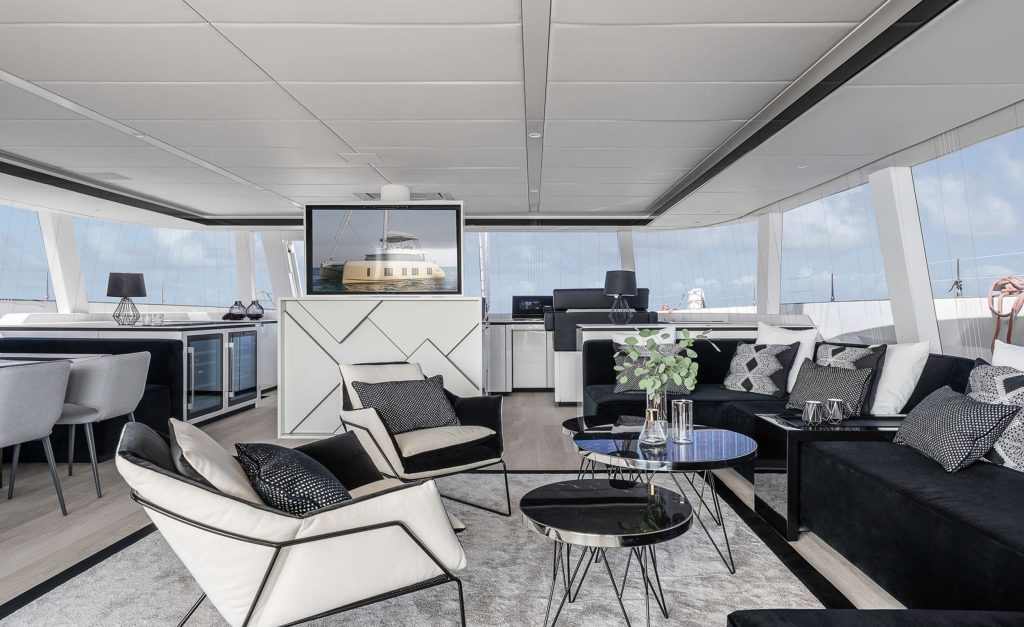 Modern yacht interior with large windows, black and white decor, lounge chairs, sofas, glass tables, and a TV displaying a boat image. Perfect for those seeking a stylish yacht charter experience with ocean views and elegant dining in the background.