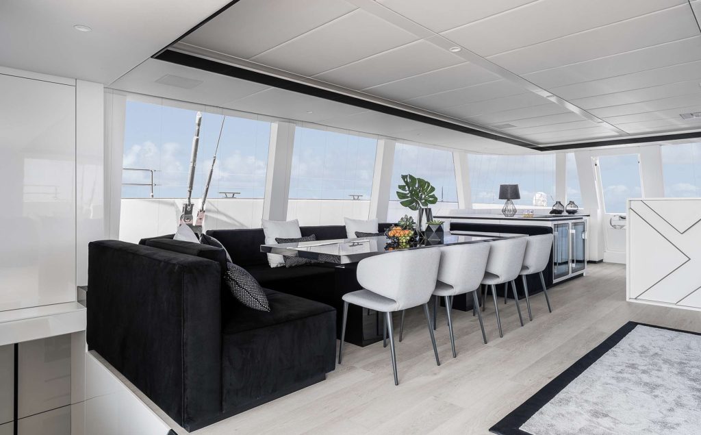 Modern yacht interior with floor-to-ceiling windows, a black sofa, white chairs around a dining table, a kitchenette, and decorative accents. Bright and airy atmosphere with ocean views—perfect for your next yacht charter experience.