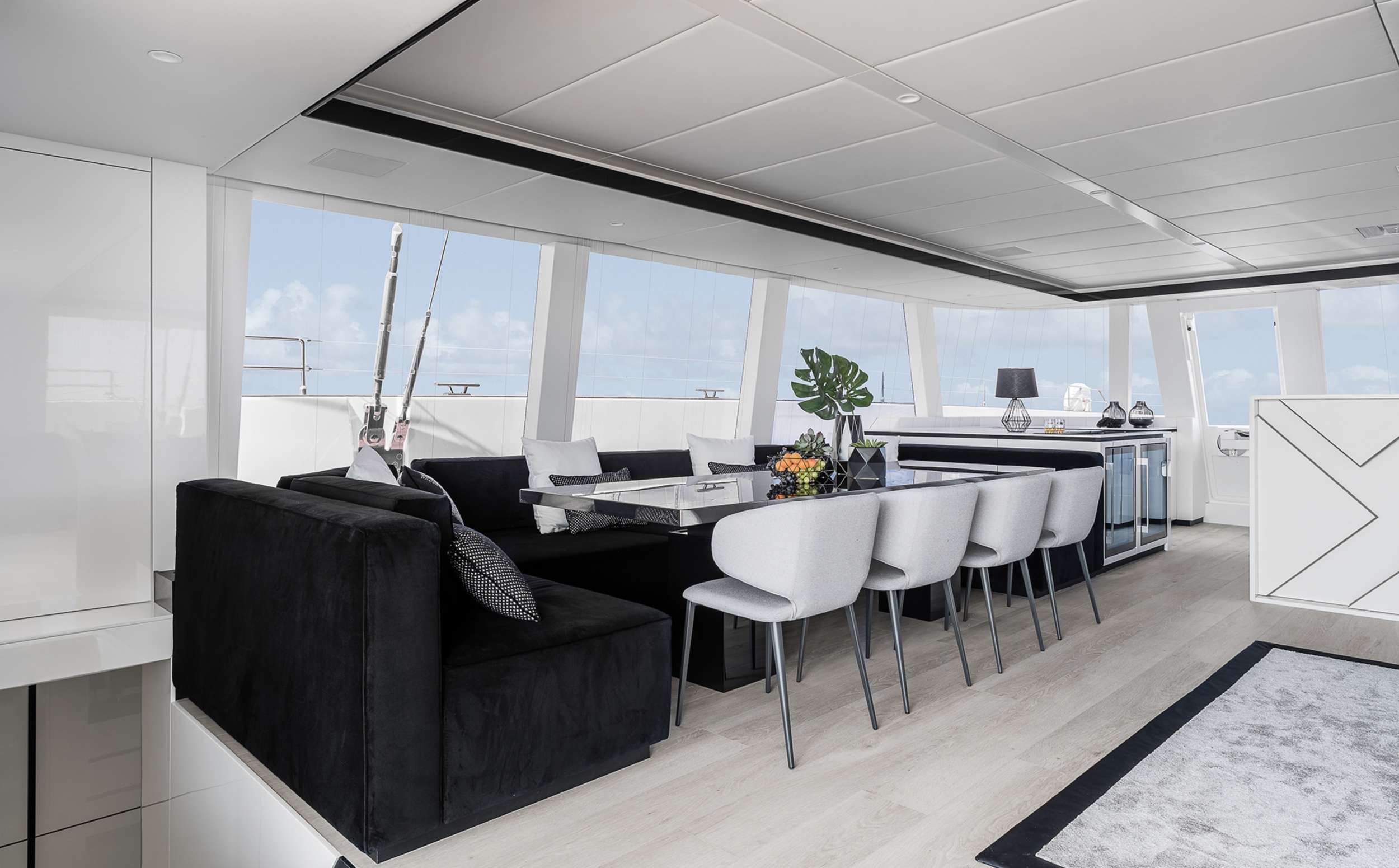 Modern yacht interior with floor-to-ceiling windows, a black sofa, white chairs around a dining table, a kitchenette, and decorative accents. Bright and airy atmosphere with ocean views—perfect for your next yacht charter experience.