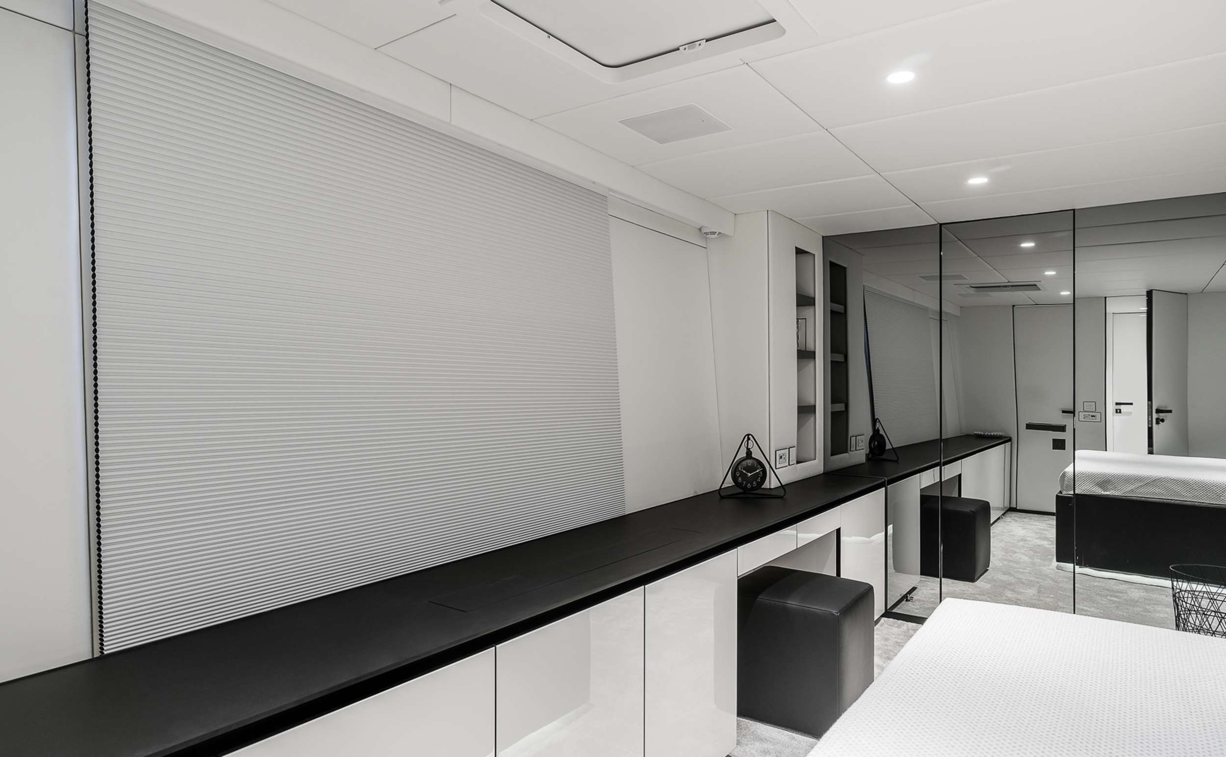 Modern minimalist bedroom with white walls, a large window shaded by a closed blind, a sleek black countertop, mirrored closet doors, and simple decor—including two black stools—echoing the refined elegance found on luxury yachts for charter.