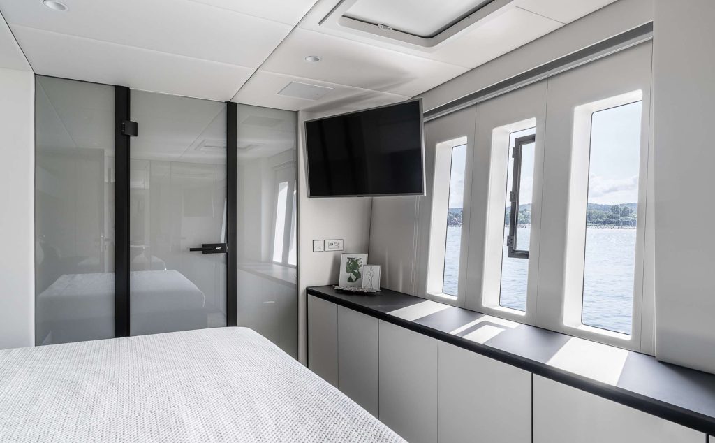 Modern bedroom with a large bed, wall-mounted TV, glass door, and wide windows overlooking the water—perfect for relaxing after a day of yacht charter. Minimalist decor features white walls and sleek cabinetry under the windows.