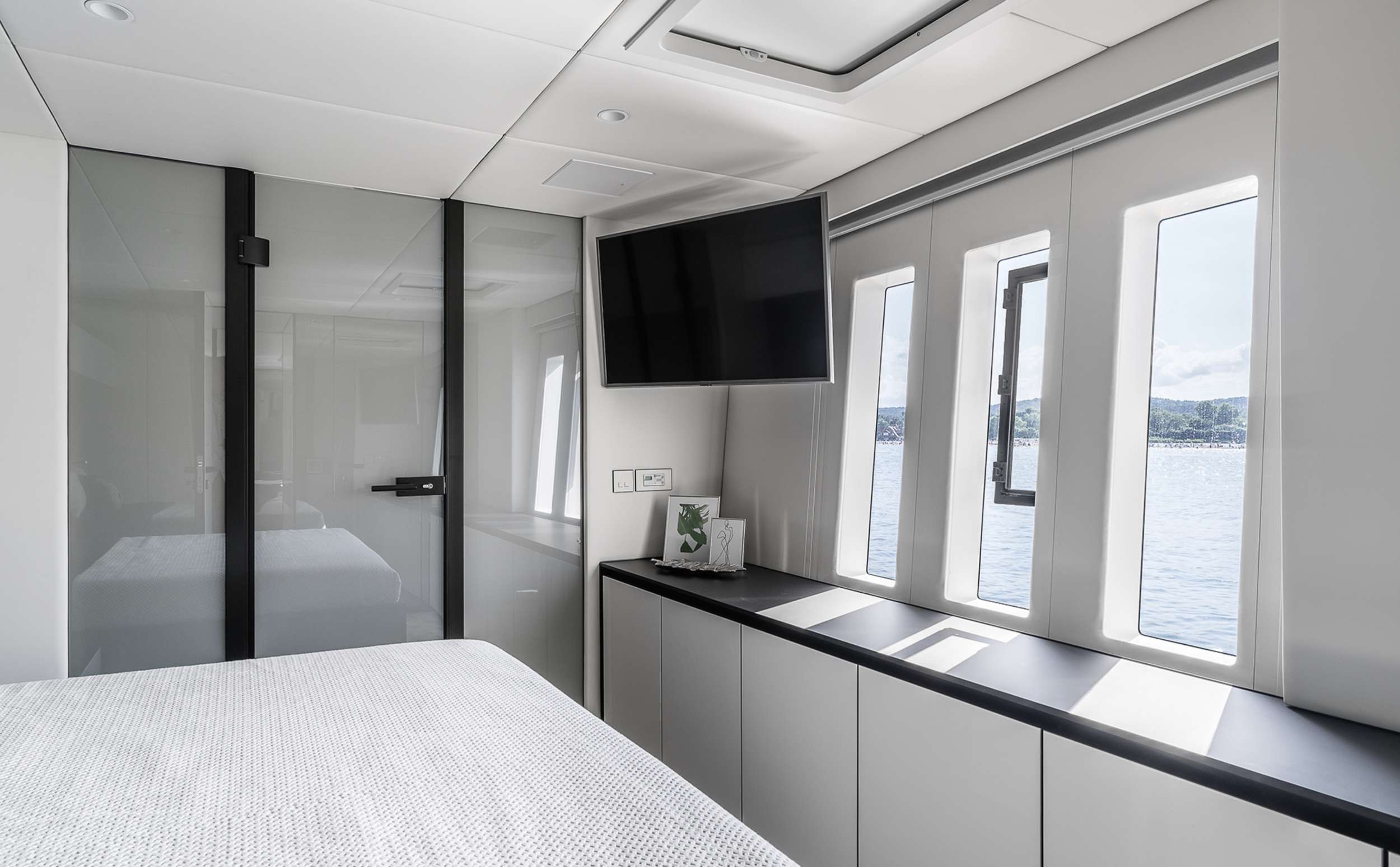 Modern bedroom with a large bed, wall-mounted TV, glass door, and wide windows overlooking the water—perfect for relaxing after a day of yacht charter. Minimalist decor features white walls and sleek cabinetry under the windows.