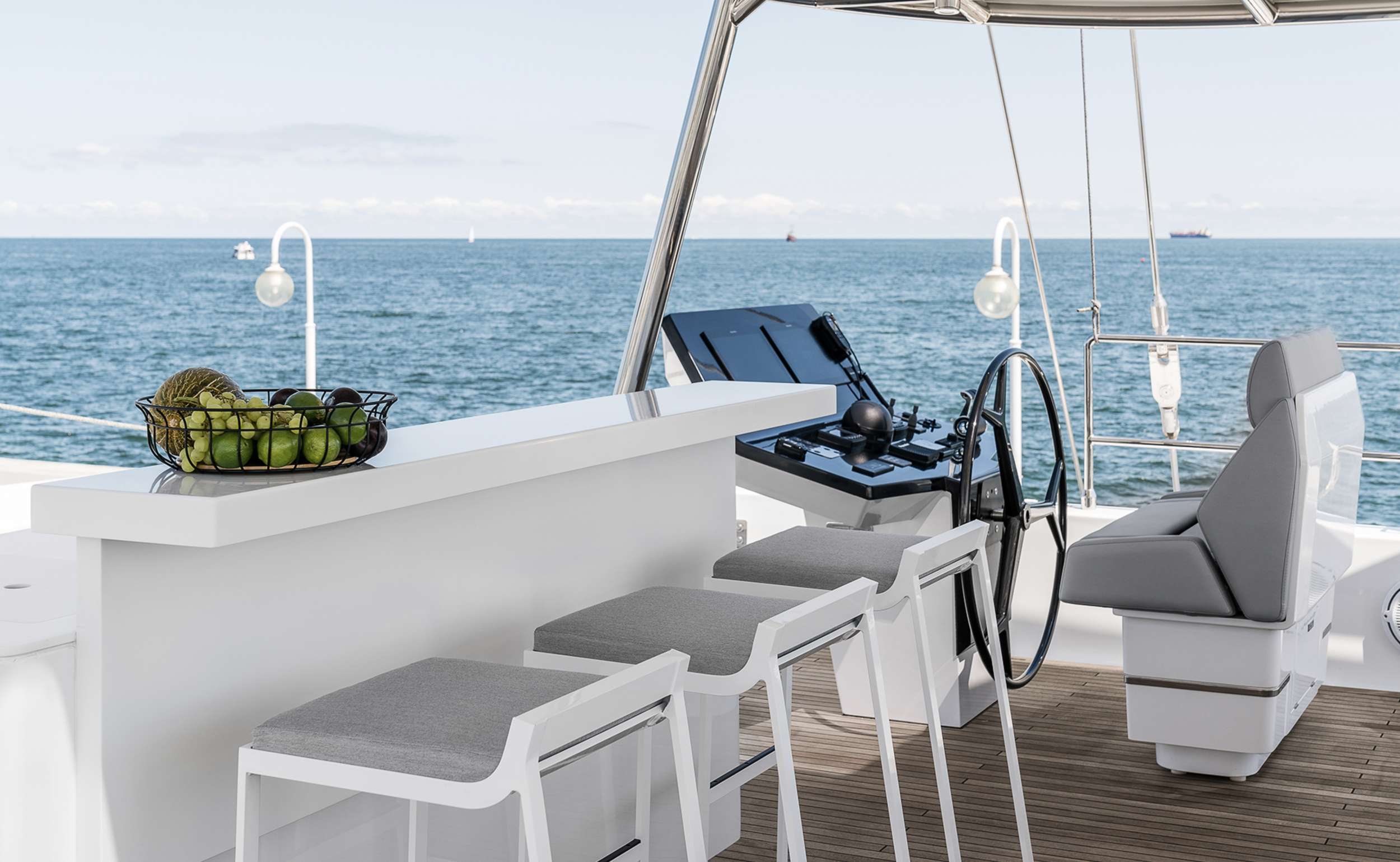 Modern yacht deck with a white bar counter, three gray-cushioned stools, a bowl of fruit, a steering wheel, and ocean views—perfect for relaxing moments on your next yacht charter under clear skies with calm waters and distant ships.