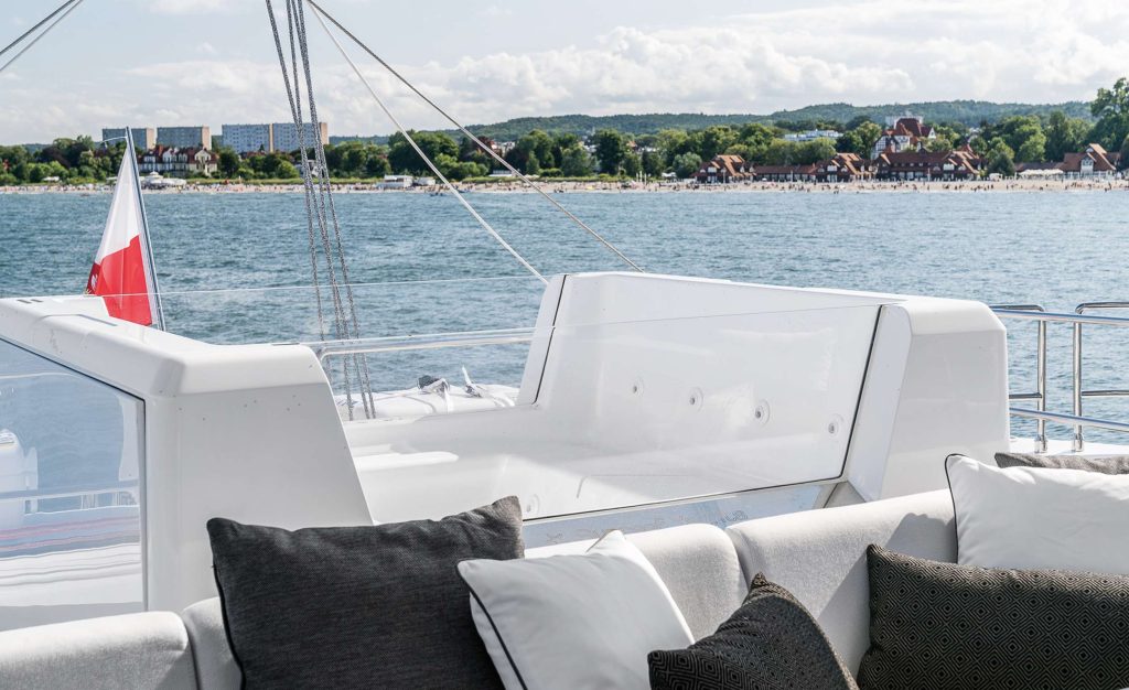 View from a yacht charter deck with white seating and dark pillows, looking out over calm water toward a distant sandy beach, green trees, and buildings along the shoreline under a partly cloudy sky.