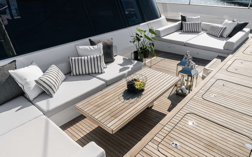 Luxurious yacht deck with white cushioned seating, striped and solid pillows, a wooden table with fruit, decorative lanterns, and potted plants—perfect for relaxing sunlight-filled moments on your next yacht charter.