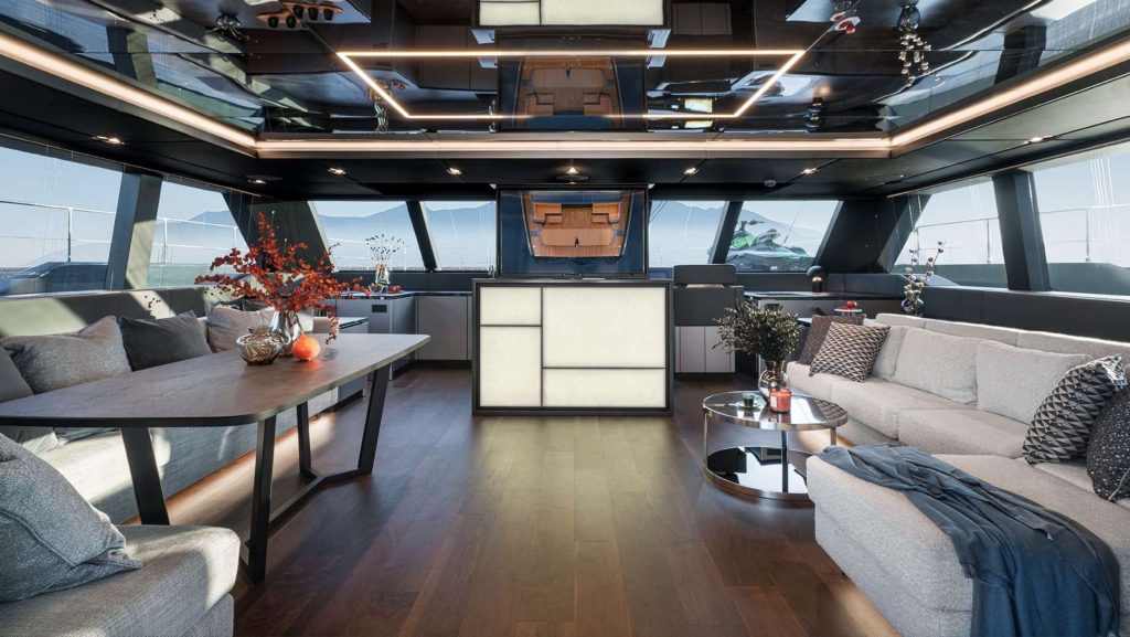Modern yacht interior featuring a spacious lounge with a gray sectional sofa, a round coffee table, dining table with chairs, decorative plants, and large windows offering ocean views—perfect for an unforgettable yacht charter experience.