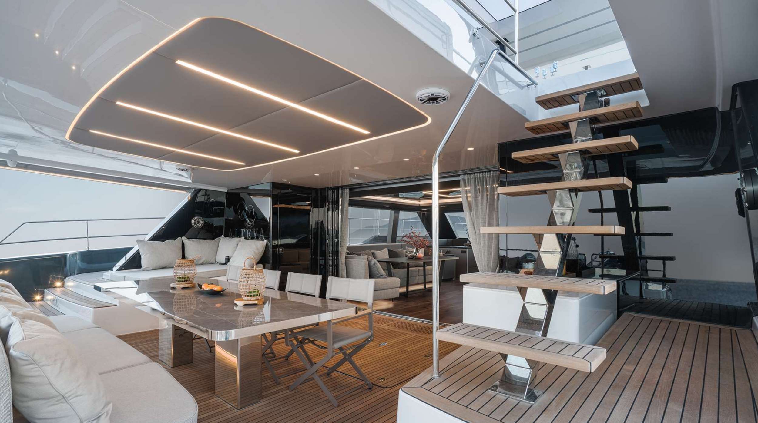 A modern yacht’s interior with a sleek dining area, cushioned seating, a glass table, and wooden steps leading to an upper level; warm lighting and large windows create an open, luxurious atmosphere perfect for yacht charter experiences.