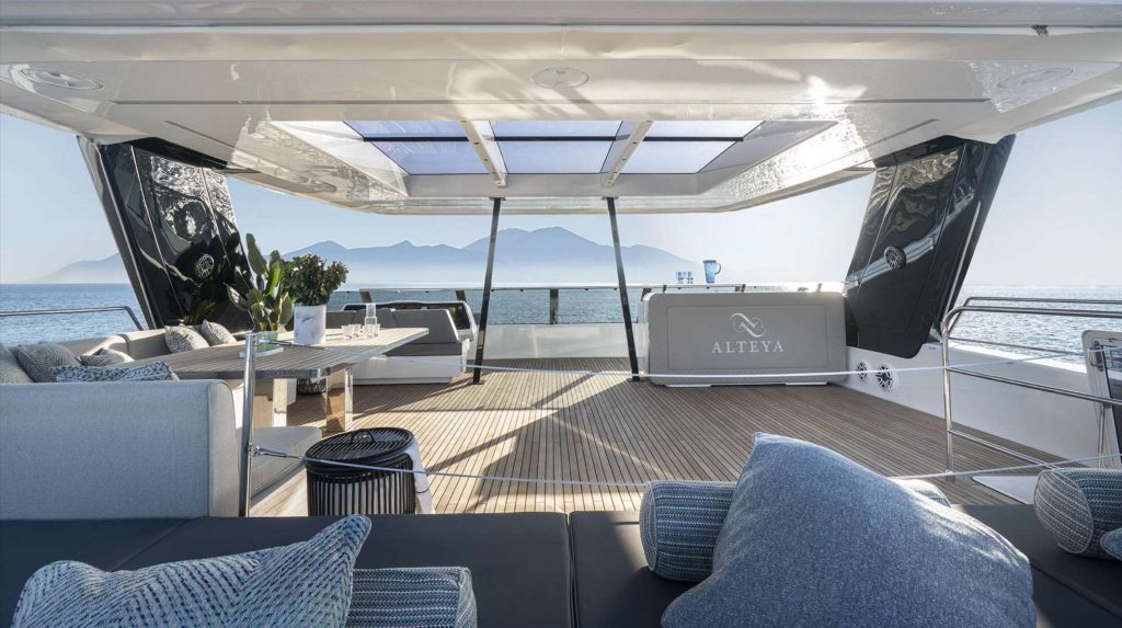 Luxurious yacht deck with modern seating, tables, and cushions, shaded by a glass roof. Perfect for a yacht charter, it offers stunning sea and mountain views beneath a clear sky. ALTEYA is written on the yacht’s back panel.