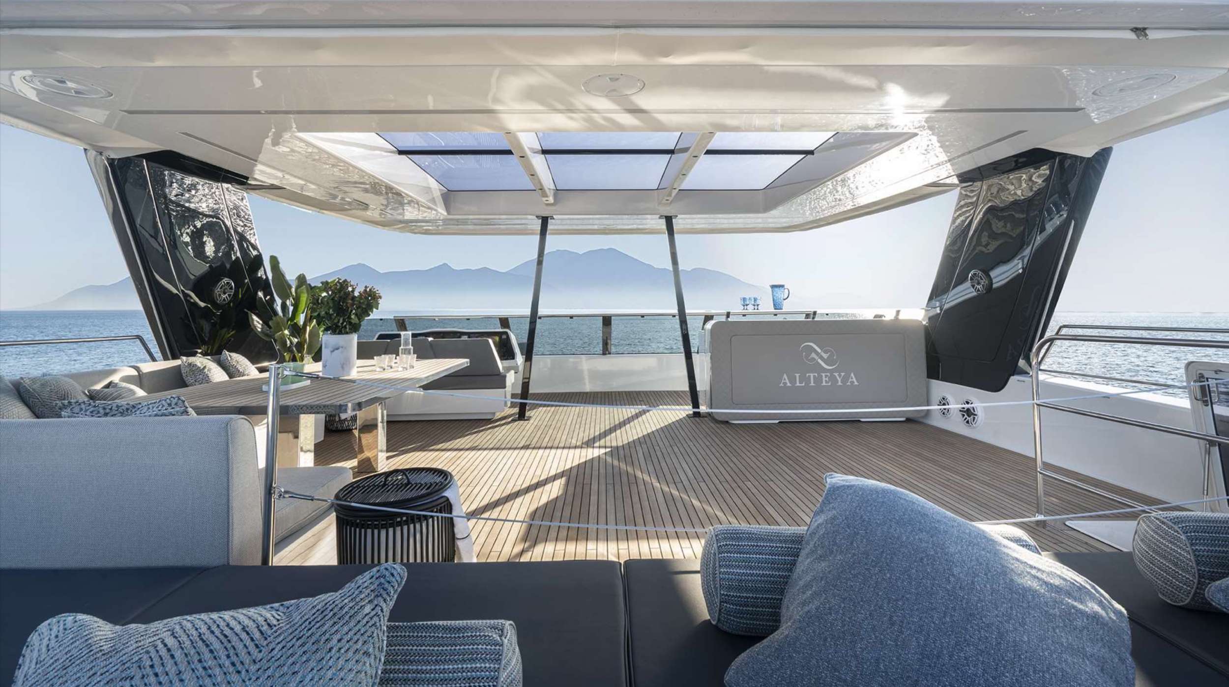 Luxurious yacht deck with modern seating, table, and plush pillows. Open roof panels let in sunlight, and there’s a view of calm sea and distant mountains. Perfect for those seeking premium yacht charter; the yacht’s name ALTEYA is visible at the back.